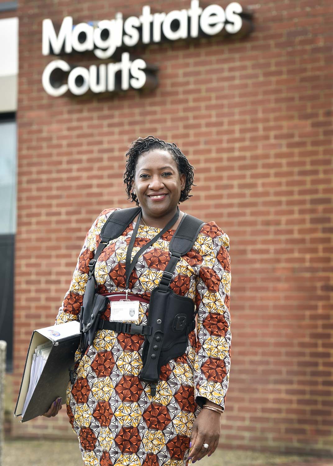 Detective Constable Yvonne Newman stands outside Magistrates Court, holding a folder and wearing a Thames Valley Police ID, and proudly wearing her Zimbawean African traditional clothing.