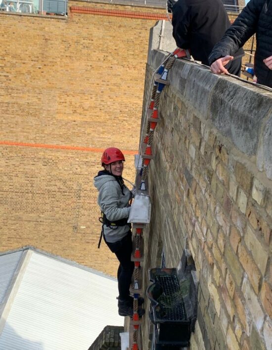 Police Sergeant Tiffany Massey scaling the side of a light brick building on a rope ladder. She is wearing a red helmet and supported by a harness. She is completing her rope ladder course, required for boarding the ships on the River Thames,