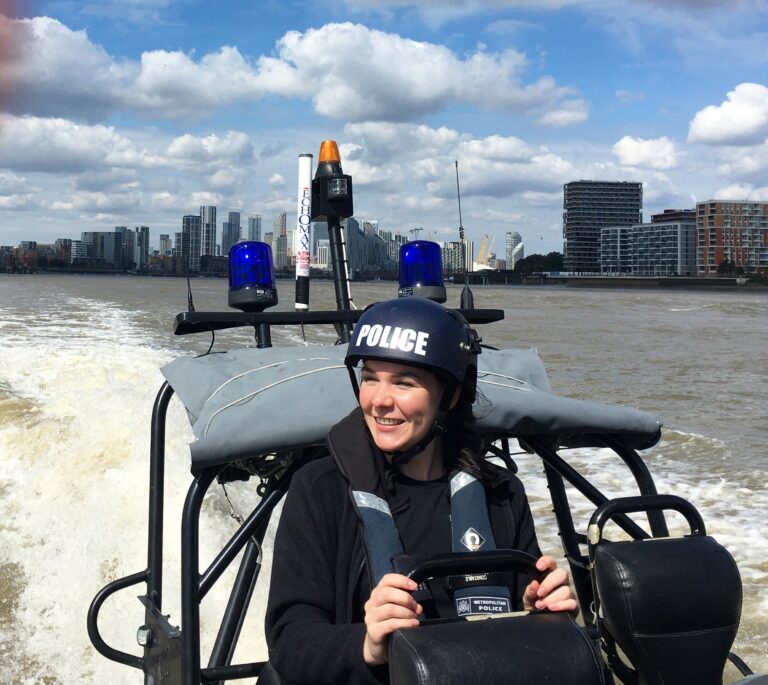 Police Sergeant Tiffany Massey sits at the back of a small black speedboat on the River Thames. The boat has two blue police lights attached and she is wearing a helmet that says 'police'. It's a sunny day and you can see the river bank behind her lined with tall London buildings.