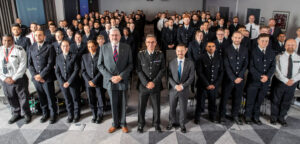 A group shot from the front of a large room, of the latest cohort of officers on Police Now's National Graduate Leadership Programme and the guest speakers at the attestation ceremony (from left to right: Justice of the Peace Clive Harper, Chief Constable Chris Haward and Police Now's Chief Executive Officer Kurtis Christoforides)