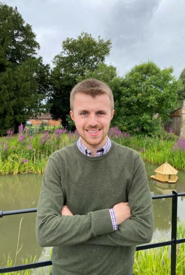 Police Constable Sam Polhill standing outdoors in front of a pond and some green trees. He's in a green jumper and smiling at the camera with his arms folded.
