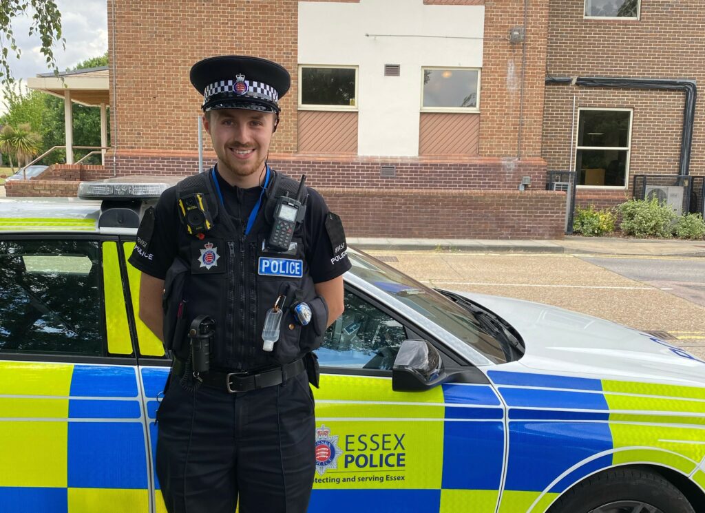 Police Constable Sam Polhill stands in front of a parked yellow and blue police car. He is smiling and facing the camera in his police uniform, outside a small block of flats.