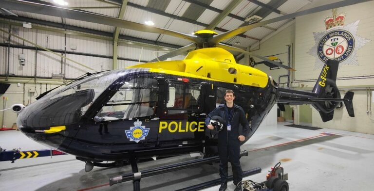 Inspector Oliver Charter is wearing a jumpsuit and standing in front of a police helicopter inside an aircraft hangar. On the wall behind, the East Midlands Air Support Unit logo can be seen.