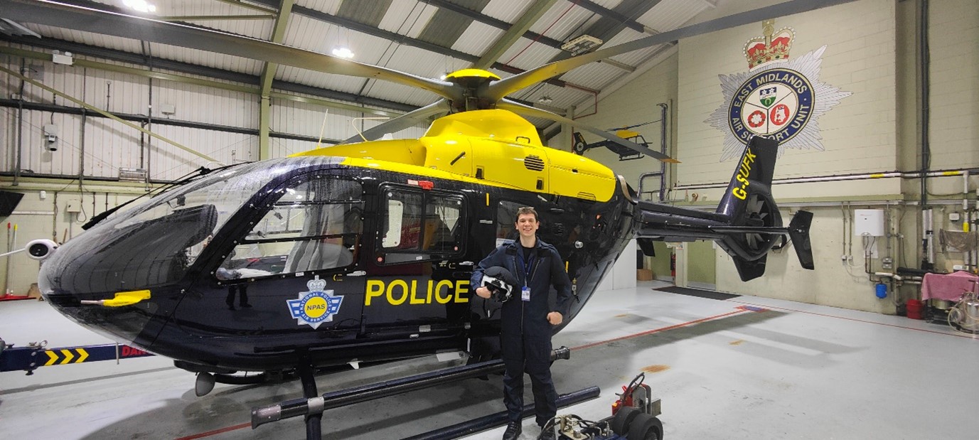 Inspector Oliver Charter is wearing a jumpsuit and standing in front of a police helicopter inside an aircraft hangar. On the wall behind, the East Midlands Air Support Unit logo can be seen.