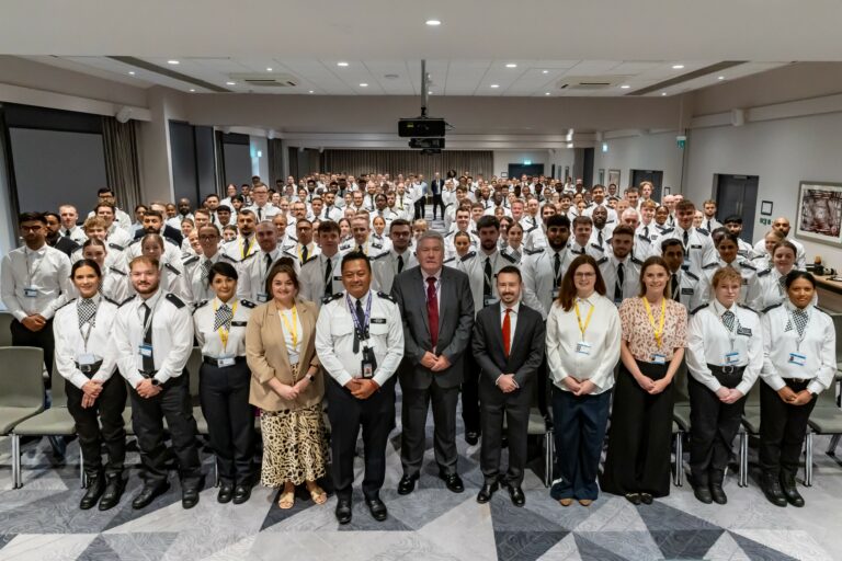A group of almost 200 uniformed officers and their academy trainers stand in rows facing the camera. At the front of the room is a line of guest speakers and Police Now colleagues.