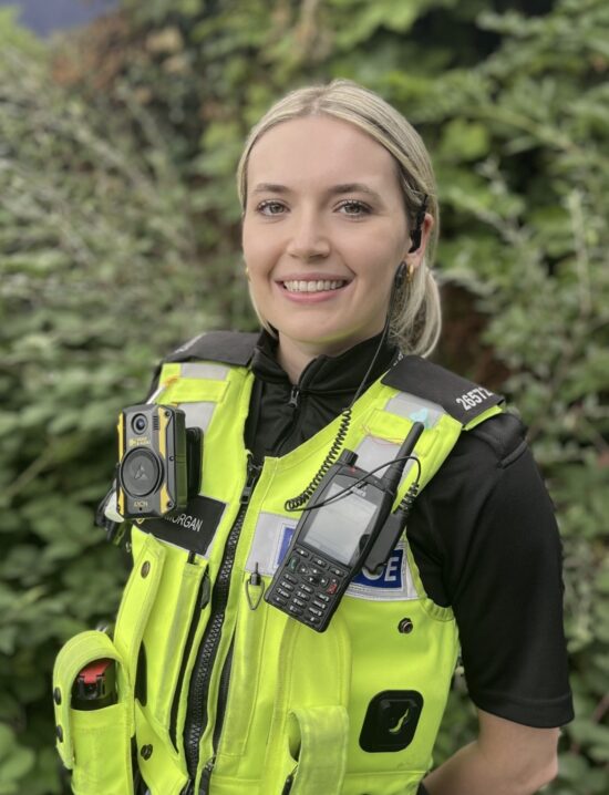 Police Constable Antonia Morgan stands in front of a green hedge in her police uniform, a yellow high vis vest prominent. She has her radio attached to her vest and is smiling at the camera.
