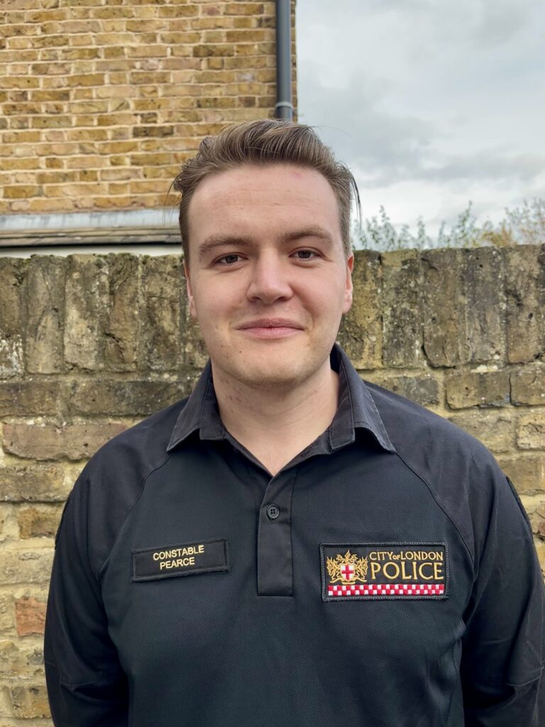 Trainee Detective Constable Blaine Pearce faces the camera, smiling and wearing a black police uniform shirt.
