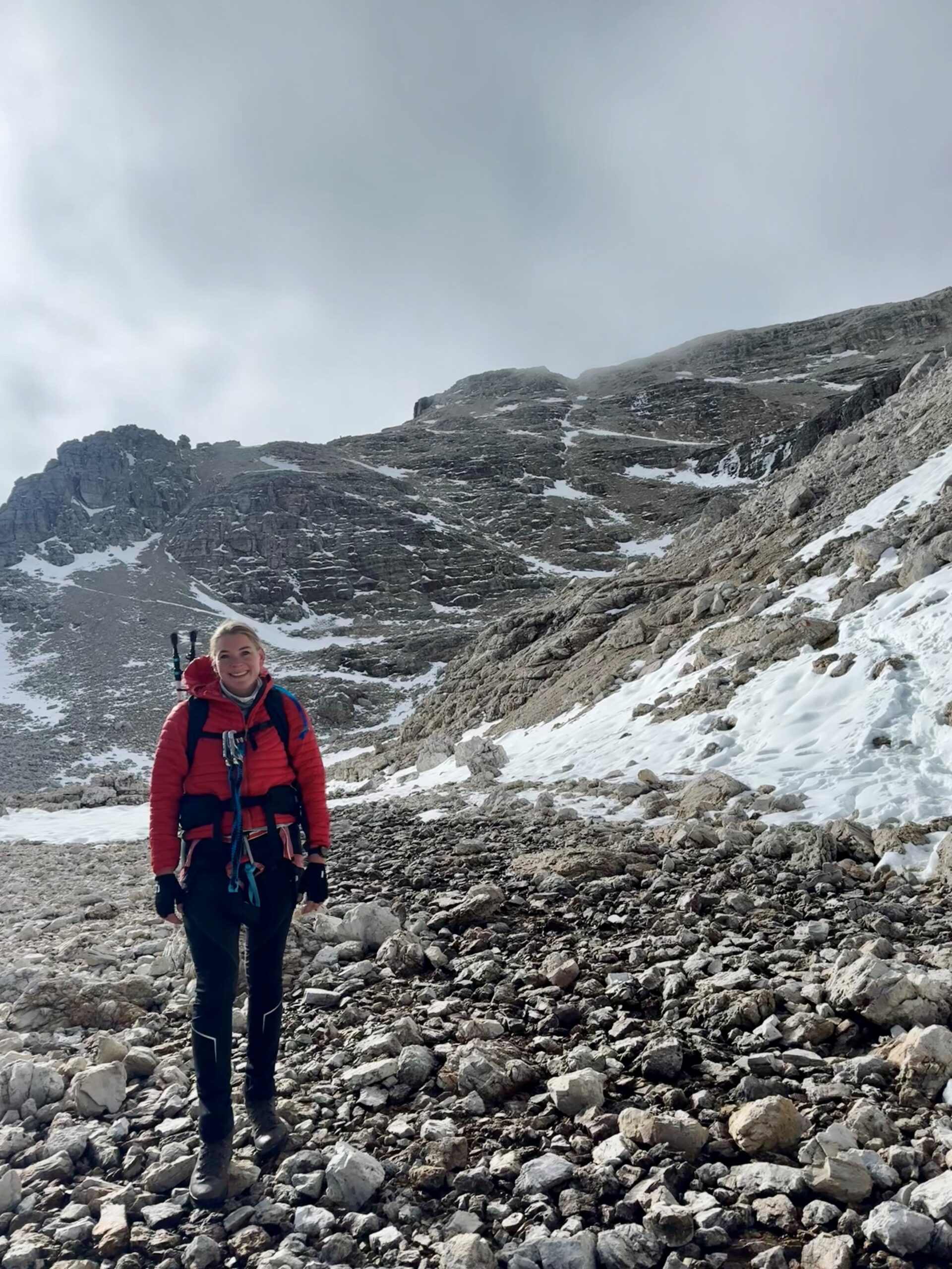 Katie Bradley stands amidst snowy hills, wearing hiking gear and wrapped in a warm red jacket - with a blue sky behind