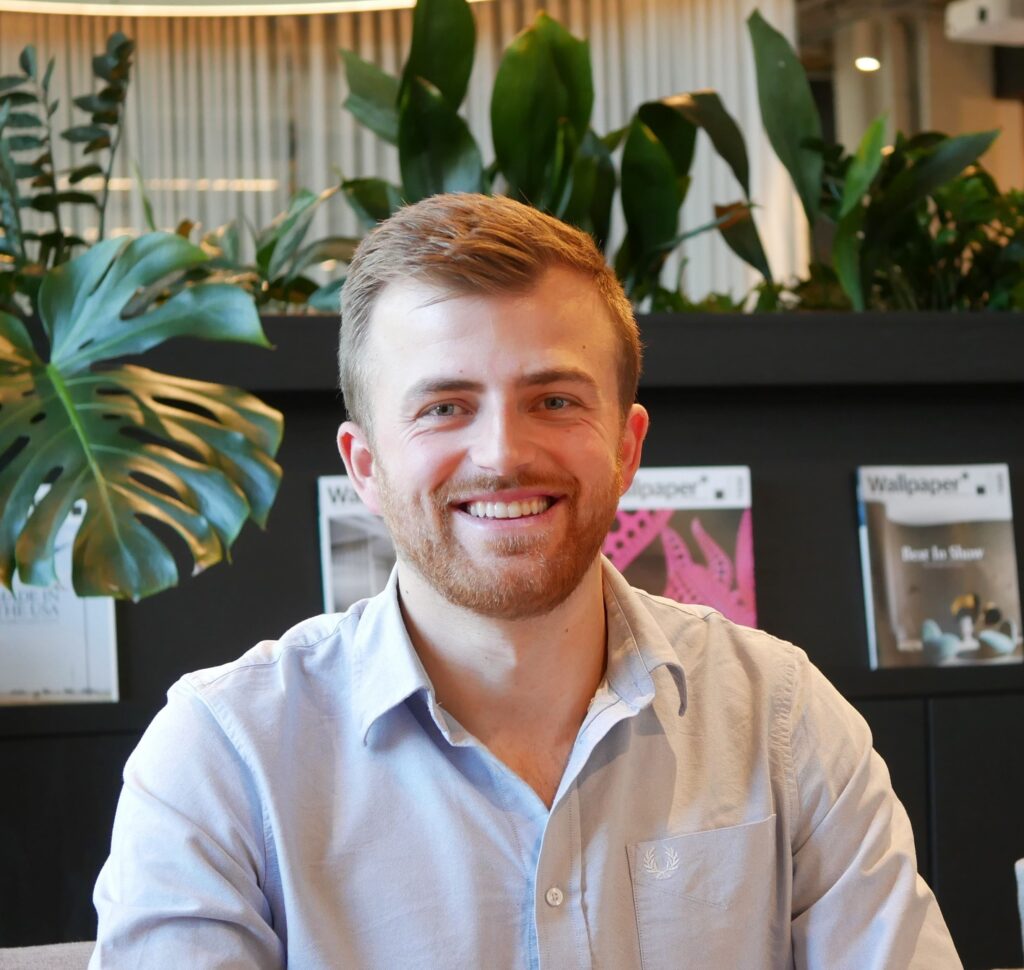 Mark Reynolds smiles at the camera. He has light brown hair and stubble and is wearing a pale, buttoned shirt.