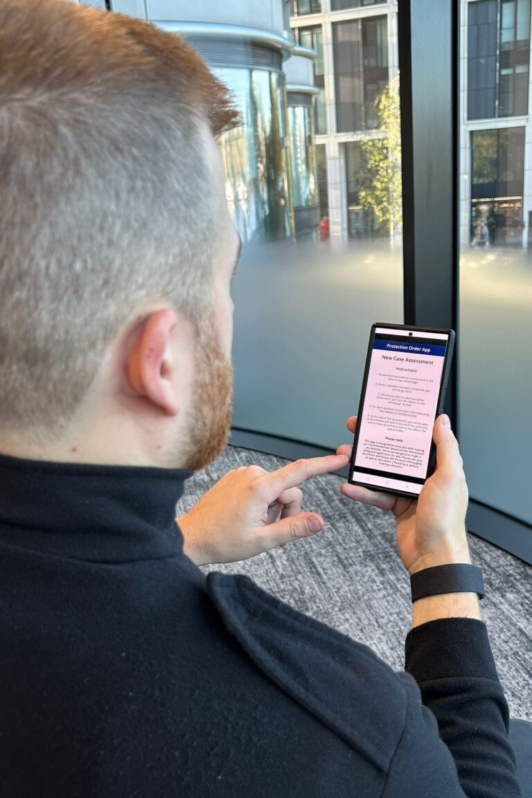 The image looks over a man's shoulder; he is holding a phone with the Protective Order App displayed