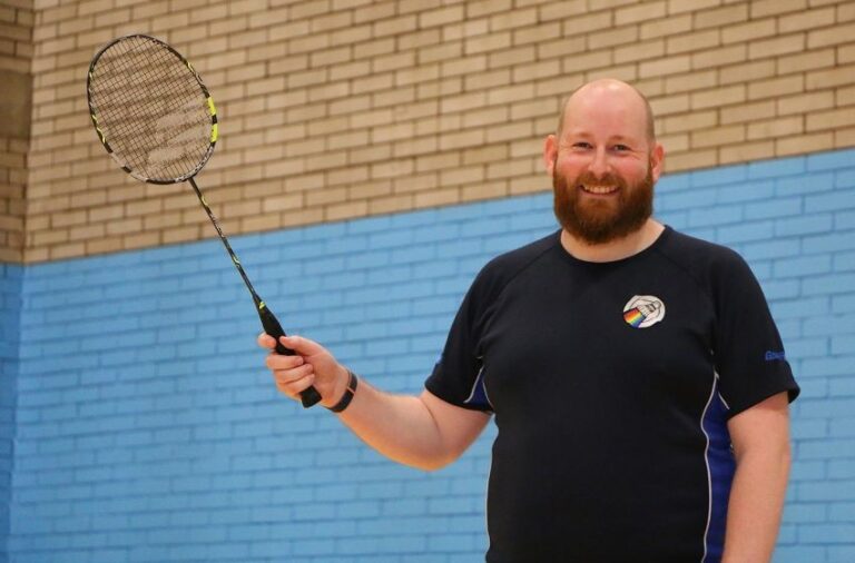 PC Peter Barnes stands in front of a brick wall on a badminton court, holding up a badminton racket. He is wearing a shirt with a rainbow-shuttlecock logo.