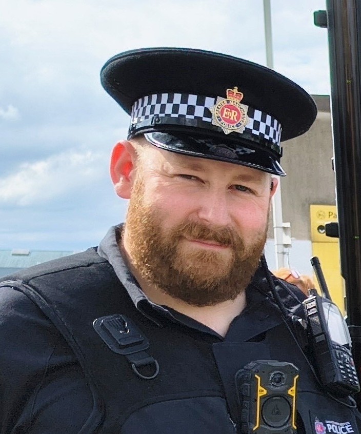 PC Peter Barnes stands in his policing uniform, smiling at the camera. He is standing outside on a sunny day.