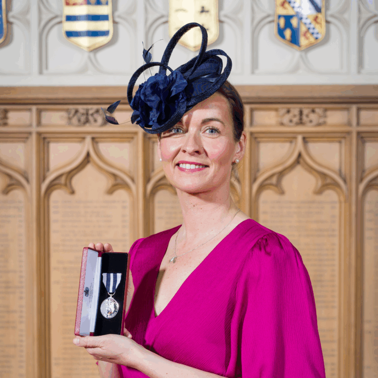Emma Harris faces the camera, holding her King's Police Medal. She is wearing formal attire and a headpiece for the Buckingham Palace ceremony.