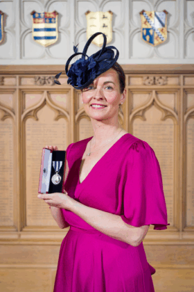 Emma Harris stands facing the camera, holding her King's Police Medal. She is wearing formal attire and a headpiece for the Buckingham Palace ceremony.