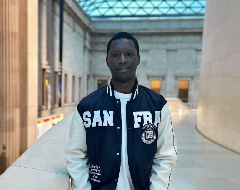 Nathaniel stands facing the camera, he is wearing a zip-up hoodie and appears to be in a grand building or museum