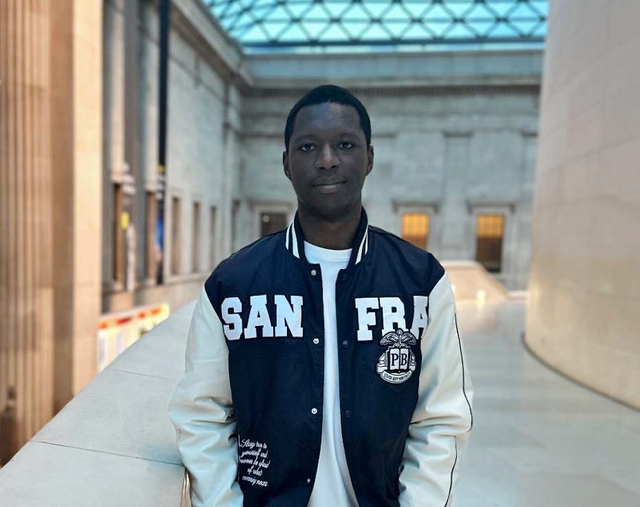 Nathaniel stands facing the camera, he is wearing a zip-up hoodie and appears to be in a grand building or museum