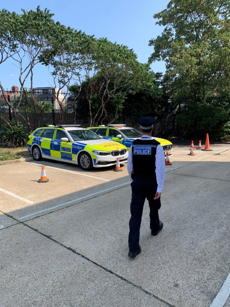 Alex strolls across a parking lot, his back to the camera. He is wearing police uniform with the words 'Metropolitan Police' on the back, and two parked police cars are nearby,