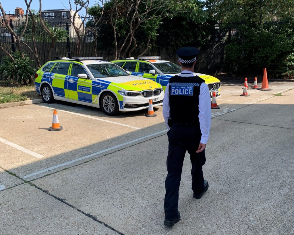 Alex strolls across a parking lot, his back to the camera. He is wearing police uniform with the words 'Metropolitan Police' on the back, and two parked police cars are nearby,