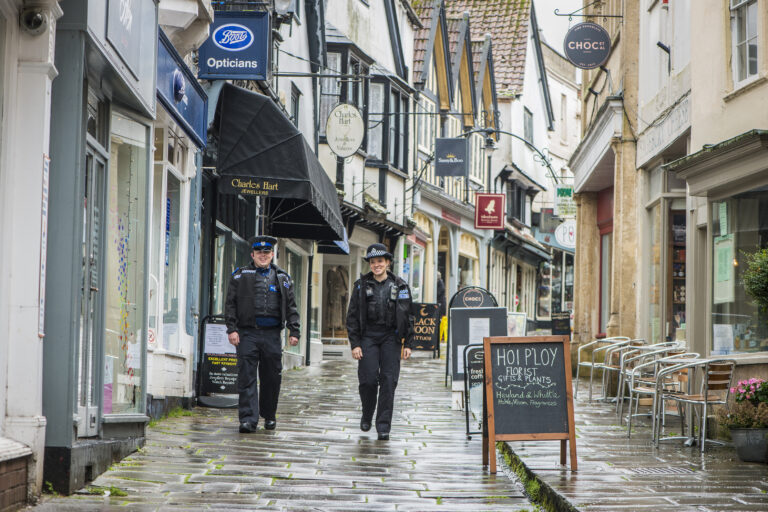 Two uniformed officers - including PC Vittoria Radaelli - are walking down a picturesque paved street, flanked by small shops. They are on patrol and smiling towards the camera.