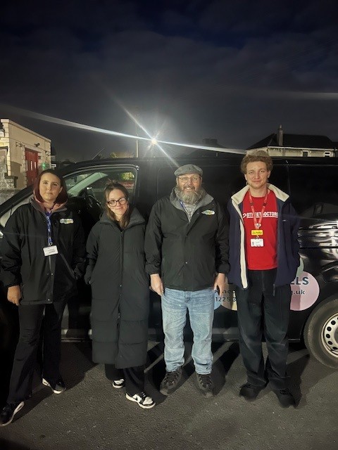A group of four stand in front of a large, black vehicle - smiling at the camera.